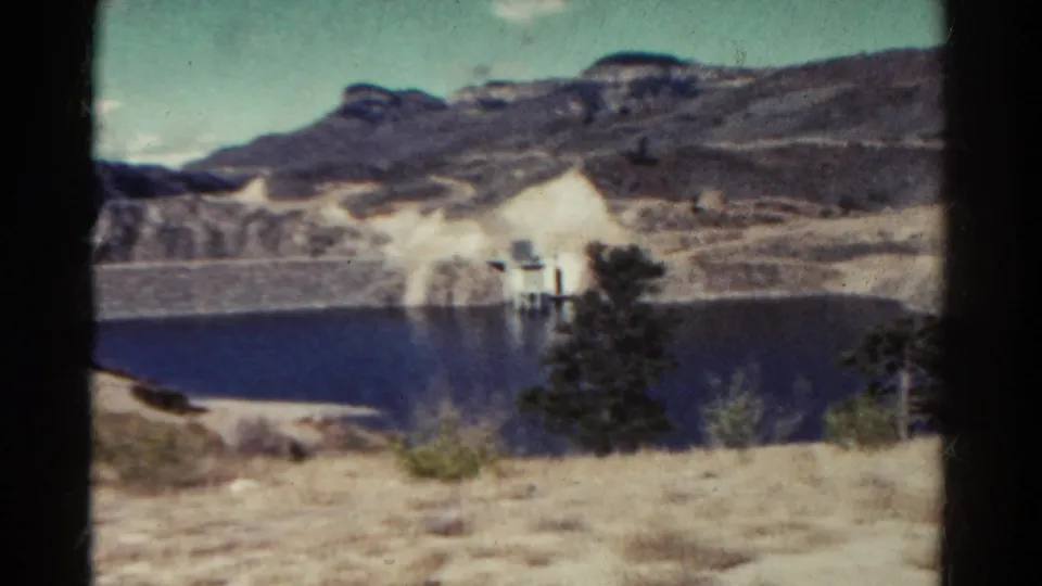 1967 Colorado home-movie view of a lake, pale rock slope, small structure, evergreens, and dry foreground terrain.