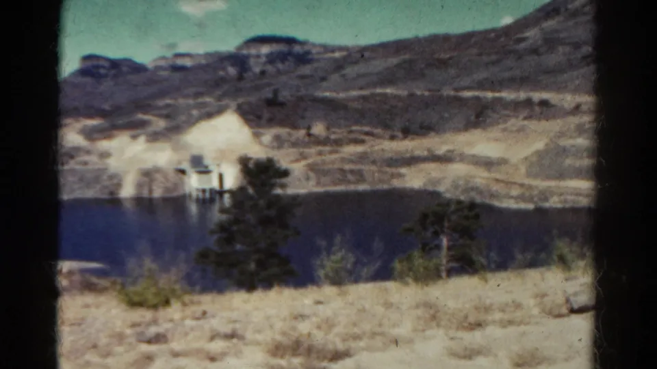 1967 Colorado 8mm frame with a small lakeside structure at left, dark water, evergreens, dry grass, and rocky terrain.