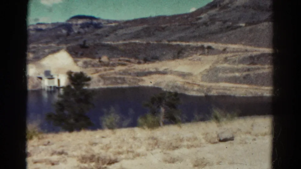 1967 Colorado mountain-lake view with the small structure near frame edge, rocky hillside, trees, dry grass, and faded 8mm color.