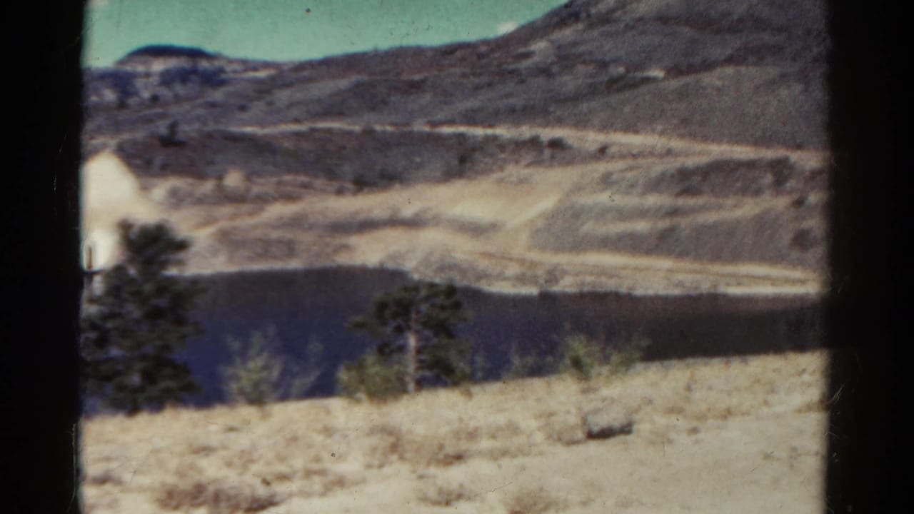 1967 Colorado 8mm frame showing a blue lake below pale dry slopes, with evergreen trees and brush near the camera.