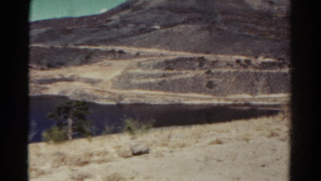 Blue lake and pale shoreline below rugged hills, framed by a dark film edge.