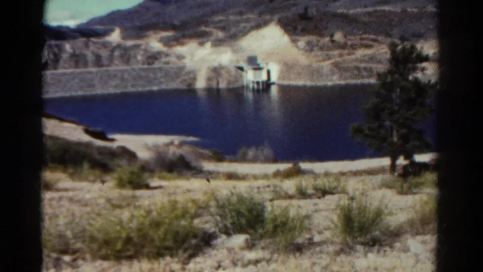 1967 Colorado 8mm frame of a lake with a small dam-like structure on the far shore, dry terrain, brush, and a tree in the foreground.