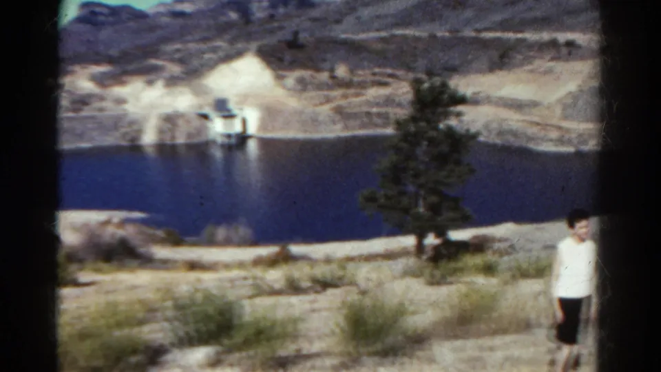 1967 Colorado 8mm frame with a standing boy or young person near the lake edge, with a tree, brush, dry hills, and a distant structure behind.