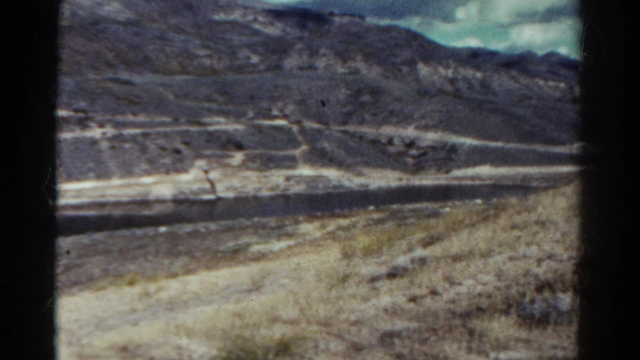 Dry Colorado hillside with distant mountain ridges, a pale road or shoreline, and cloudy sky.