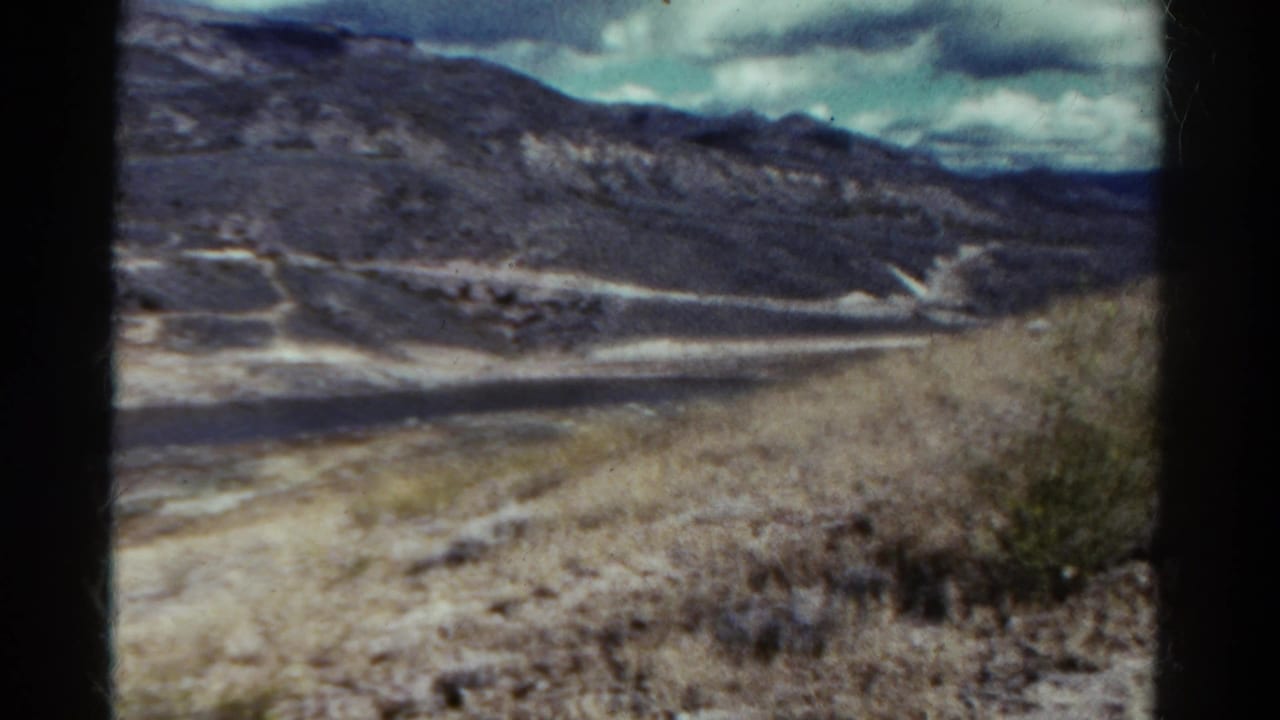 1967 Colorado travel frame with a dry slope in the foreground and a winding pale road or shoreline below distant mountains.