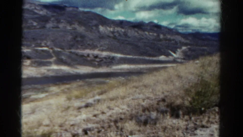 1967 Colorado travel frame with a dry slope in the foreground and a winding pale road or shoreline below distant mountains.