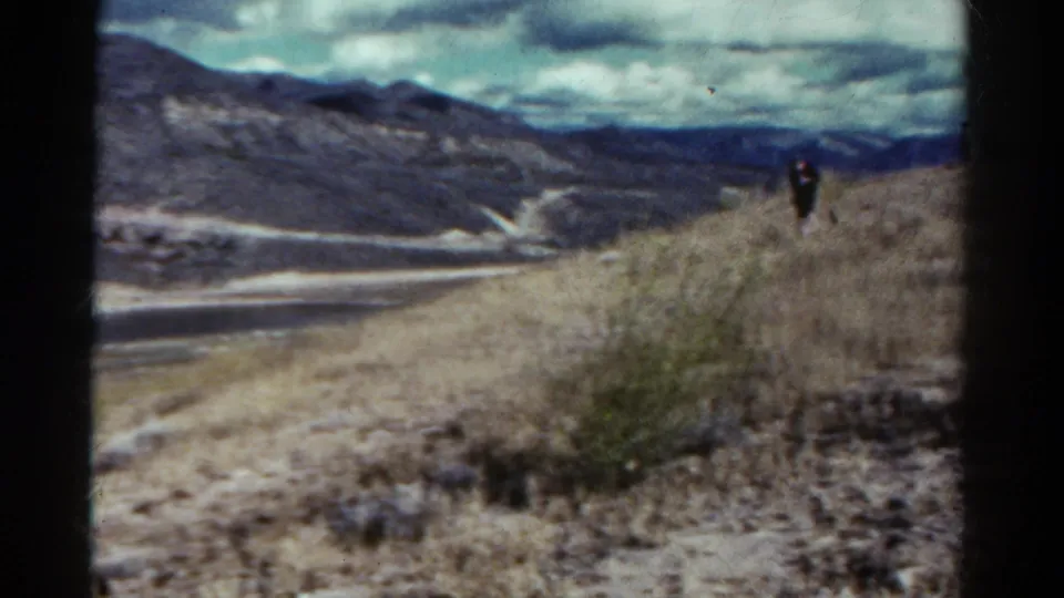 1967 Colorado 8mm frame showing a small distant figure walking on a dusty trail above rugged hills and a pale route below.