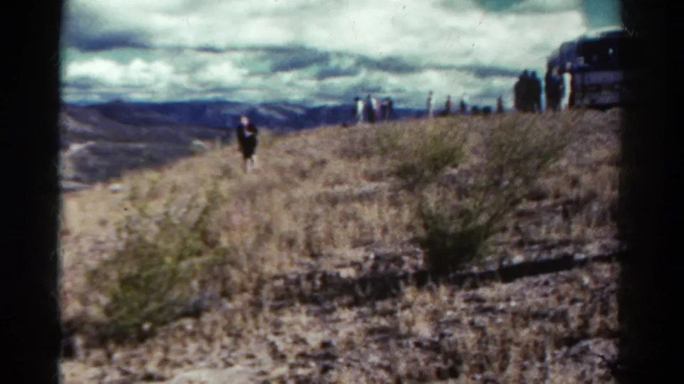 1967 Colorado home-movie frame with distant people on a dry hillside trail and a parked vehicle partly visible at the right edge.
