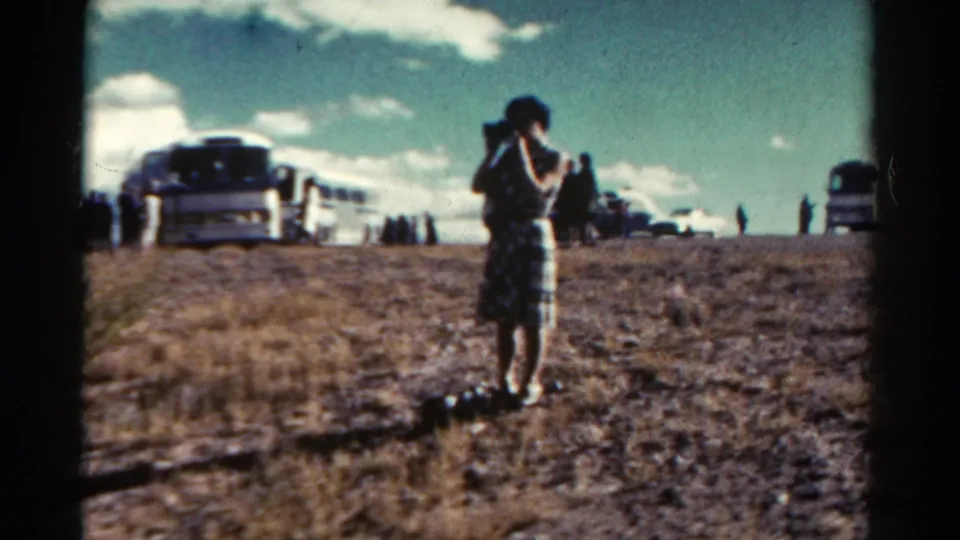 The handheld frame centers a woman in a plaid dress with a tour bus, parked vehicles, and visitors spread across the dusty Colorado overlook.