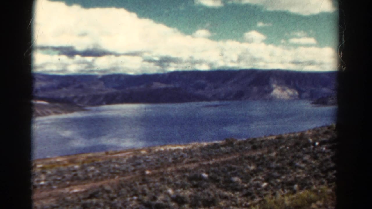 Lake and distant ridges seen beyond a rocky slope with clouds overhead.