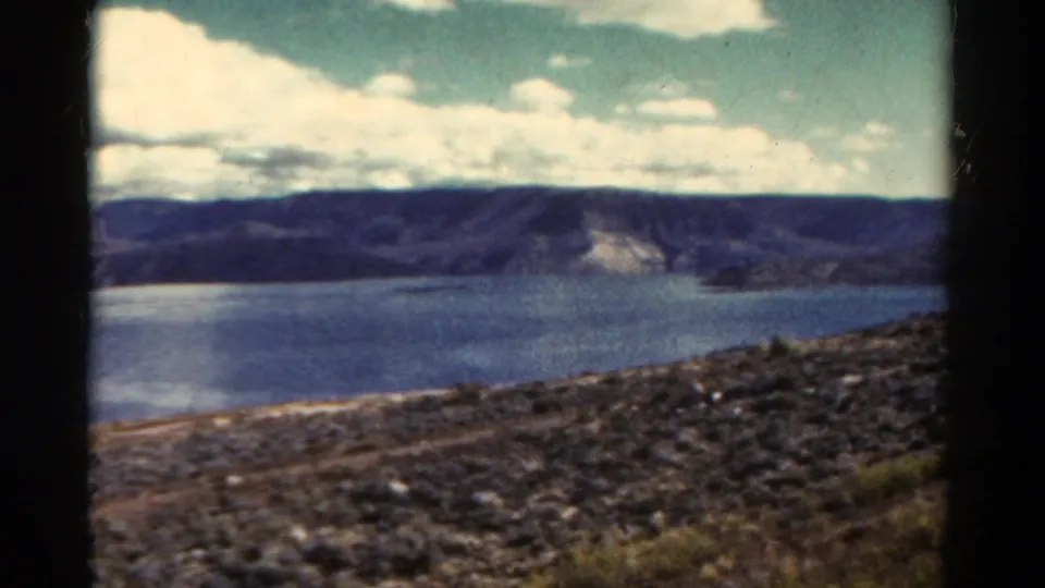 1967 Colorado 8mm still showing a mountain lake with rocky near ground, blue water, and a partly cloudy sky.