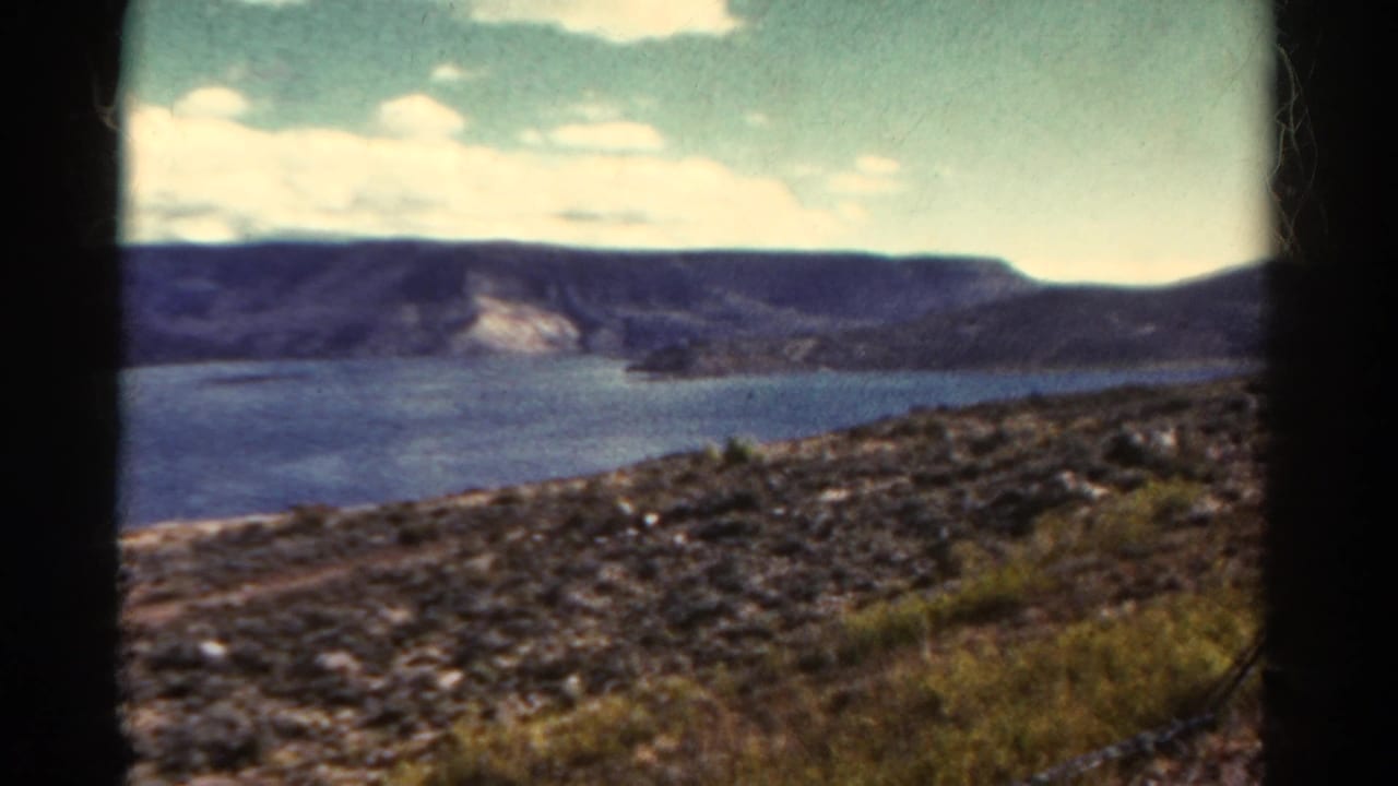 Blue lake below dark mountain ridges with rocky grass in the foreground.