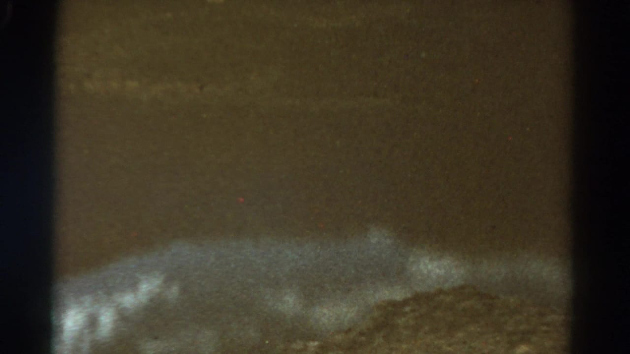 Grainy dark 8mm frame with calm water, pale sky, and a low distant hillside.
