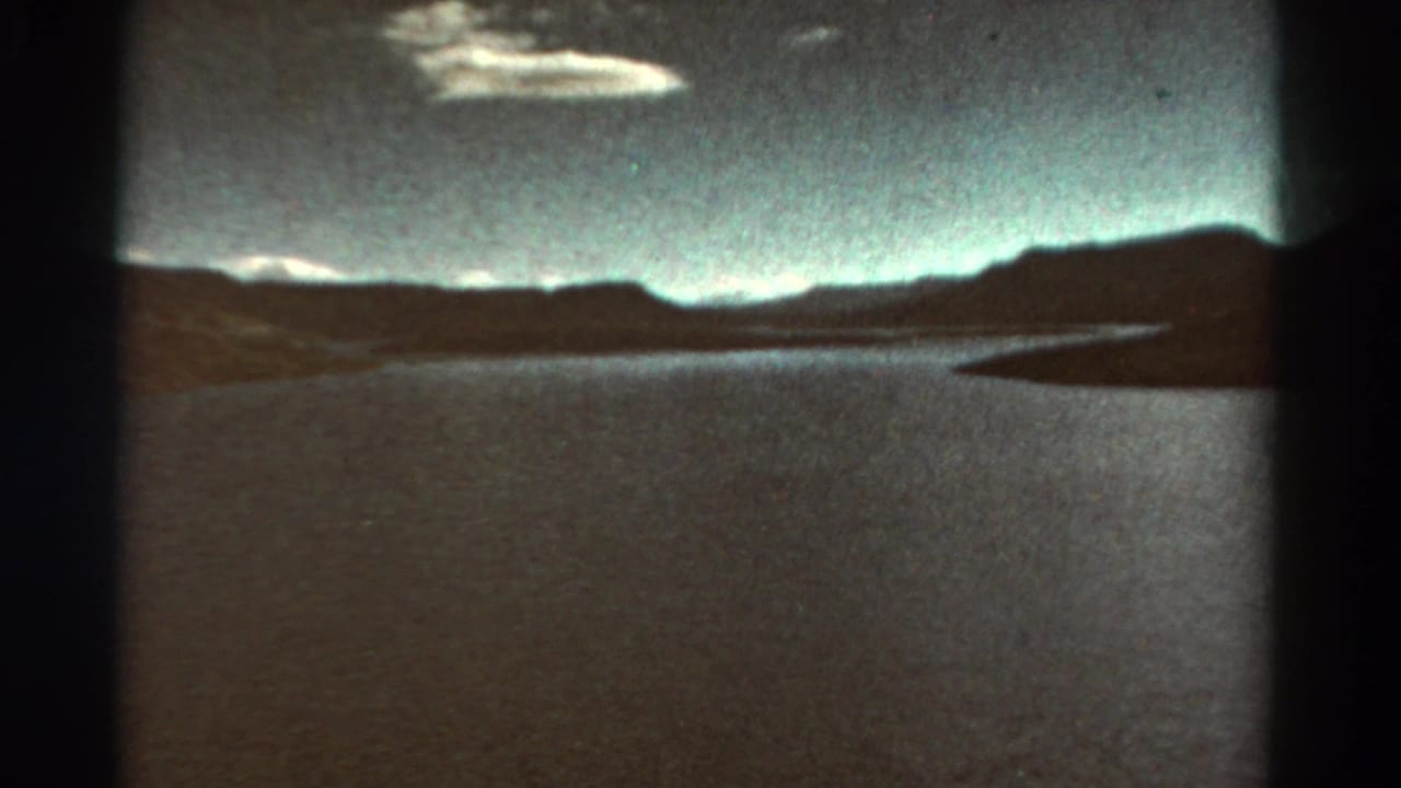 Wide waterway beneath dark mountain ridges and pale clouds, seen through rounded film-frame edges.
