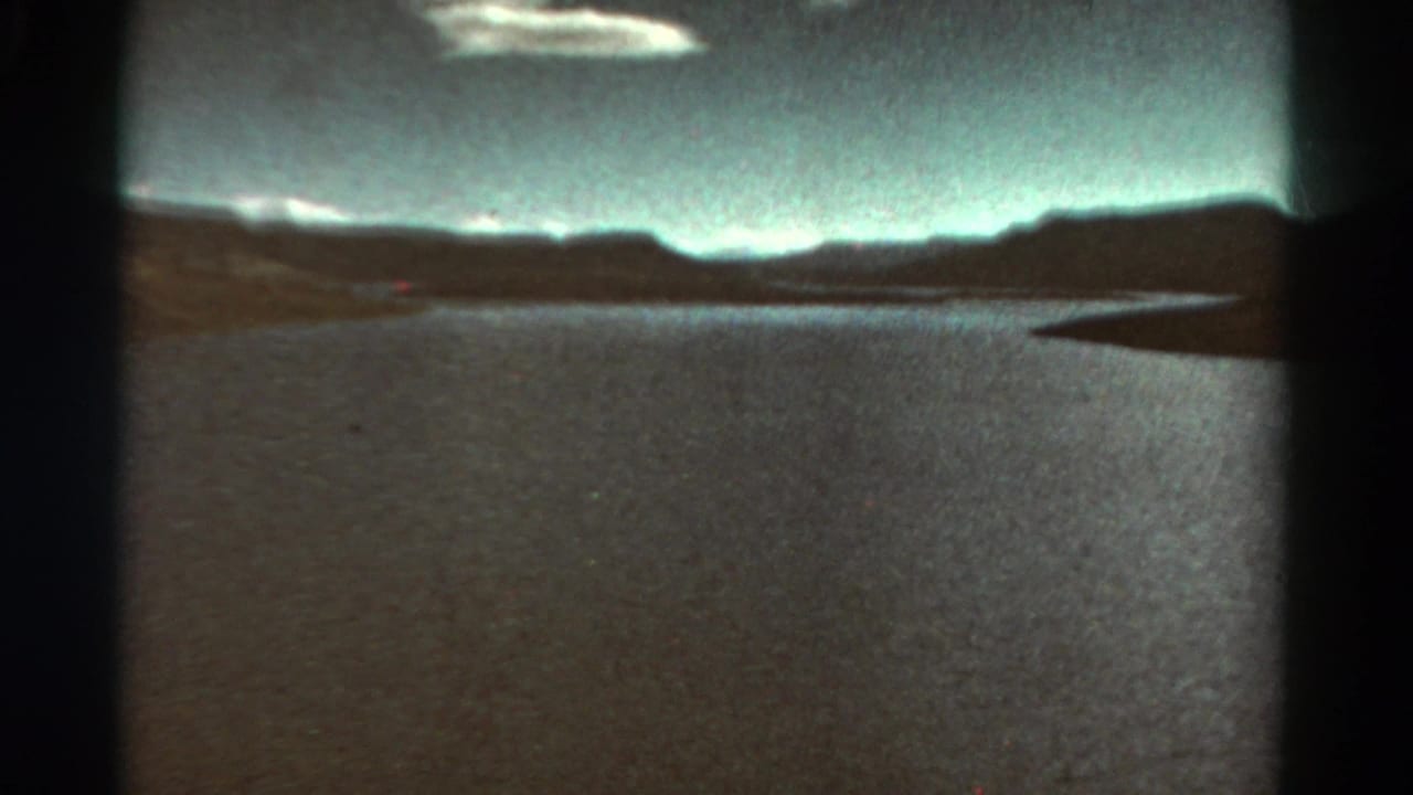 Broad water surface with distant hills, pale sky, and a dark shoreline at the right edge.