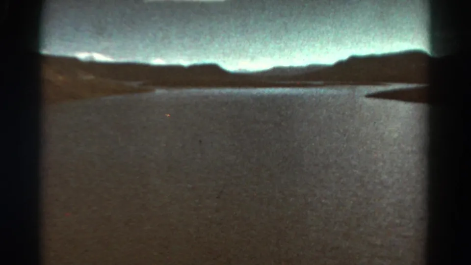 1967 Colorado home-movie frame of a quiet river or reservoir framed by hills, clouds, and rounded 8mm projection edges.