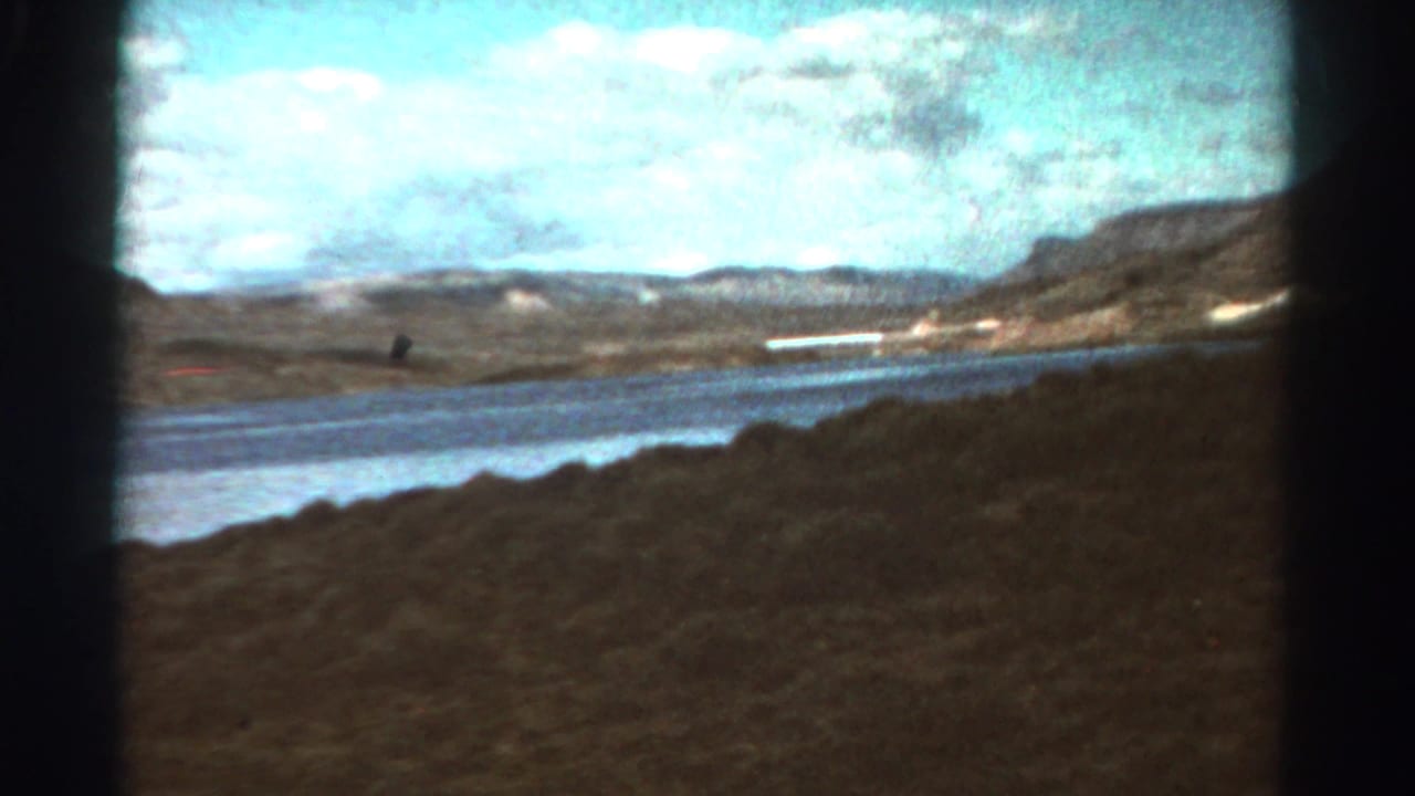 Lake and distant mountains seen from a moving roadside viewpoint under a partly cloudy sky.