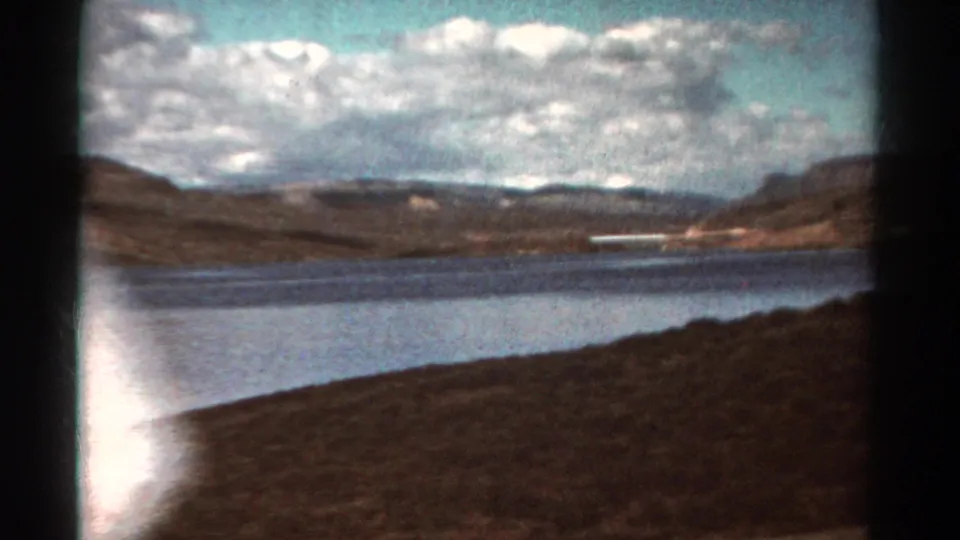1967 Colorado 8mm frame showing lake water, distant hills, cloud cover, roadside foreground, and a bright edge flare from the film scan.