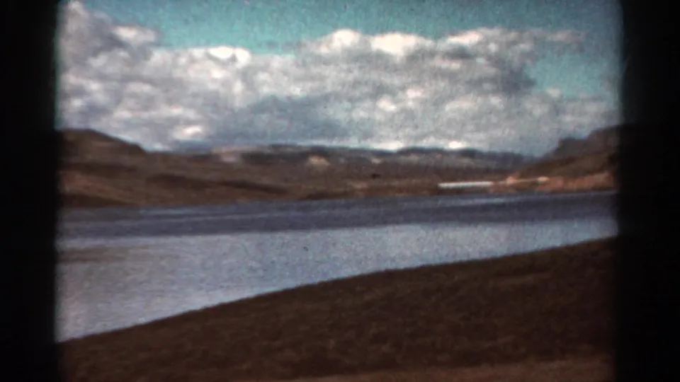 1967 Colorado home-movie frame looking over a lake toward rugged hills, with a brown roadside slope and soft 8mm color texture.