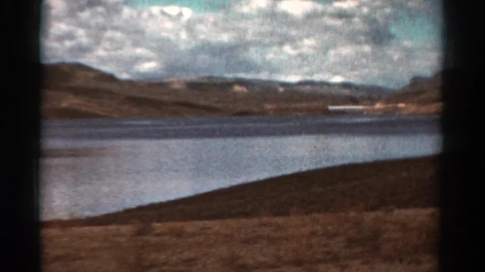 1967 Colorado lakeside frame showing water, dry hills, clouds, and a distant white structure along the shore.