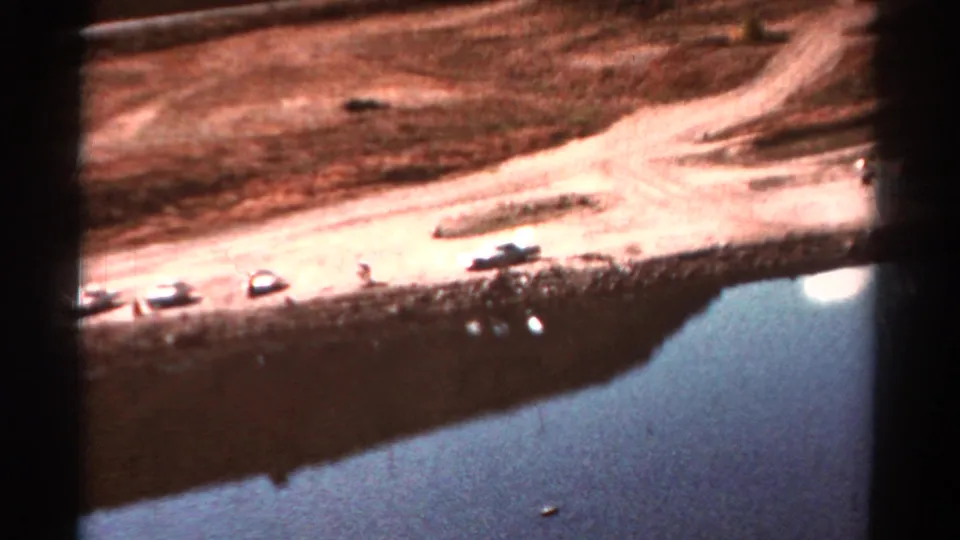 1967 Colorado home-movie frame of a lakeside dirt road, parked cars, dry ground, and water at the edge of the frame.