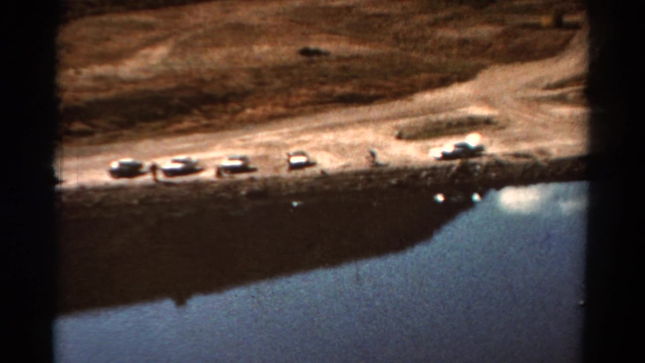 Parked cars line a dirt lakeshore above blue water with dark film borders.
