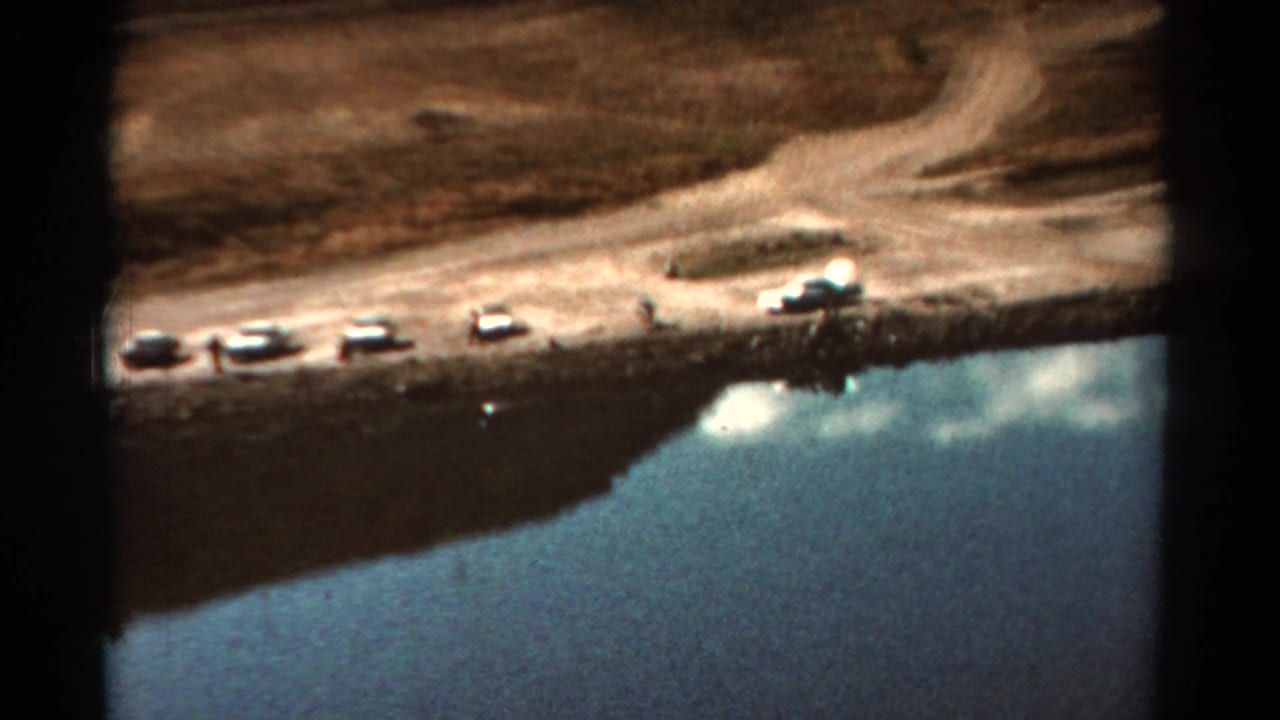 Cars and small figures appear along a dirt shore beside reflective lake water.