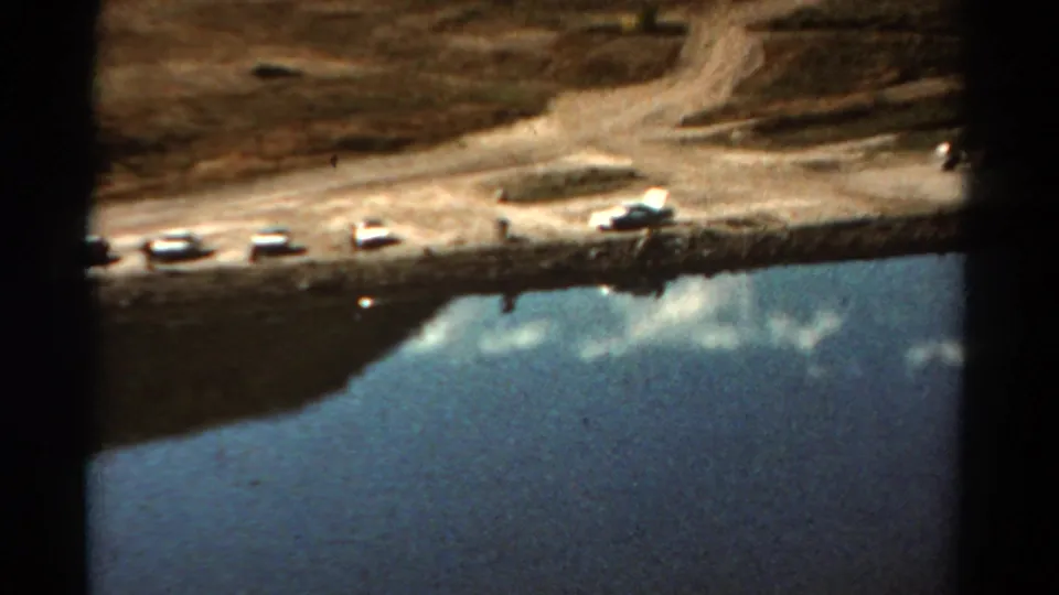 1967 Colorado 8mm still with a shoreline road, parked cars, distant visitors, and reflected sky in the lake.