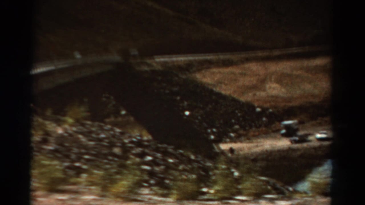 Mountain roadside view with arid slope, drainage channel, and a parked or passing vehicle in the distance.