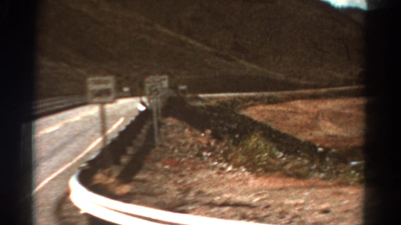 1967 Colorado mountain-road frame with a curving paved lane, metal guardrail, small road signs, dry shoulder, and brown hillside.