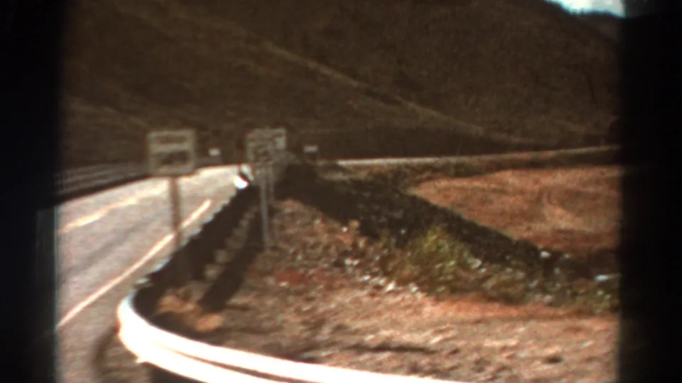 1967 Colorado mountain-road frame with a curving paved lane, metal guardrail, small road signs, dry shoulder, and brown hillside.