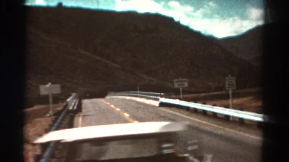 Vehicle-view 8mm frame from Colorado in 1967, looking over a car hood toward a guarded bridge or raised roadway with roadside signs.