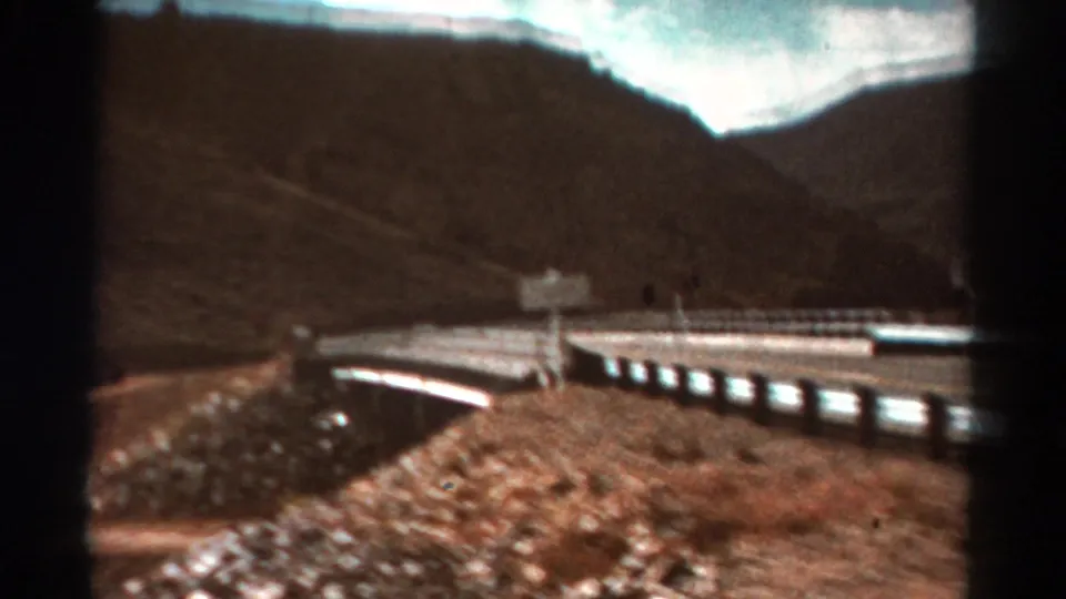 1967 Colorado 8mm frame showing guardrails and a bridge-like road structure over dry rocky terrain below dark mountain slopes.