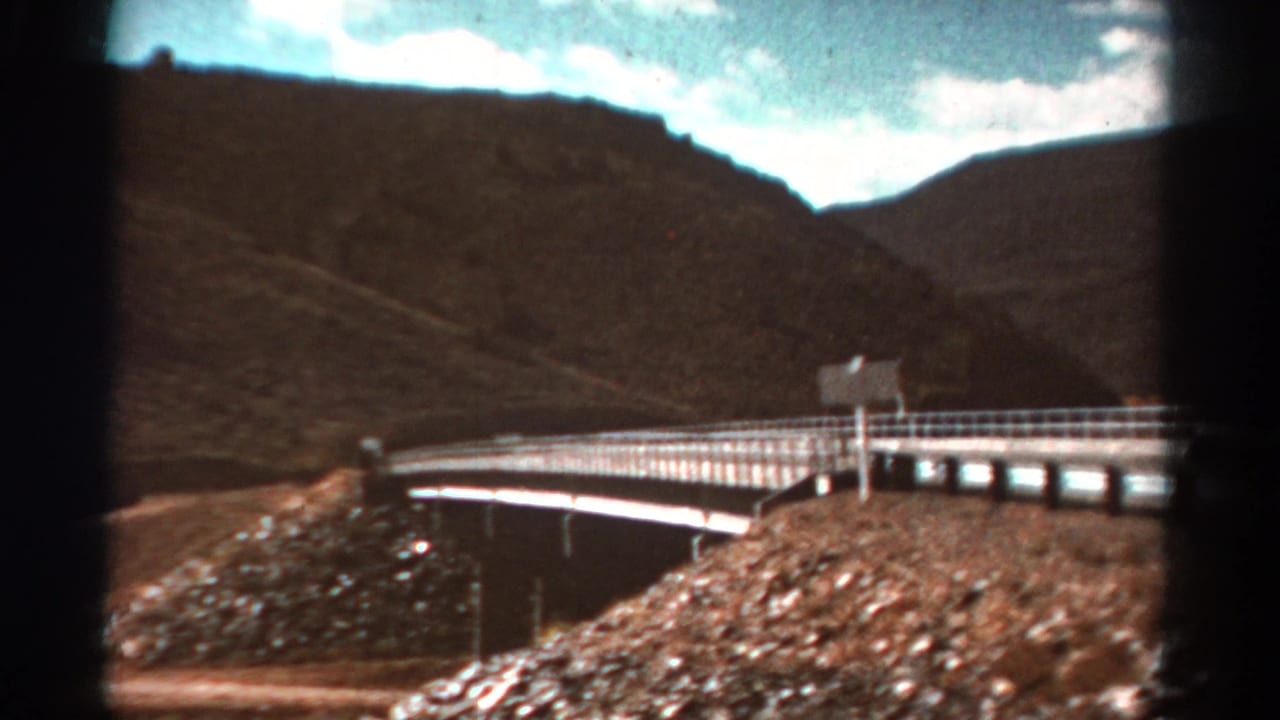 Bridge crossing a rocky riverbed below dark mountain slopes.