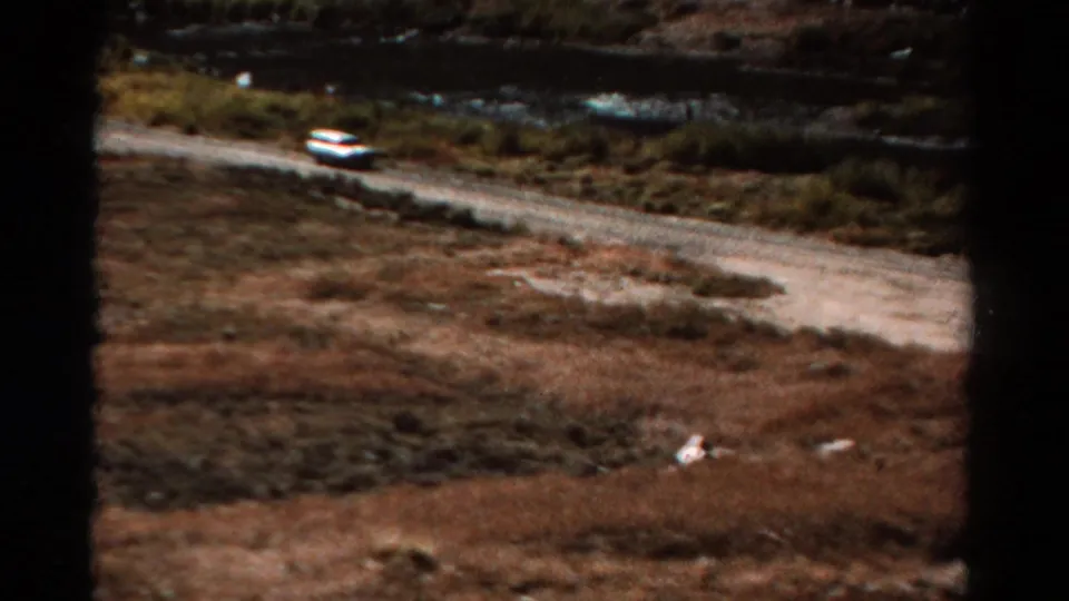1967 Colorado 8mm frame with a small car on a dirt road beside a river in rugged terrain.