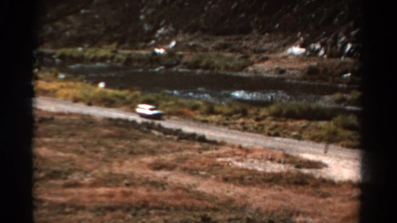 White car on a dirt road beside water and rocky hills.