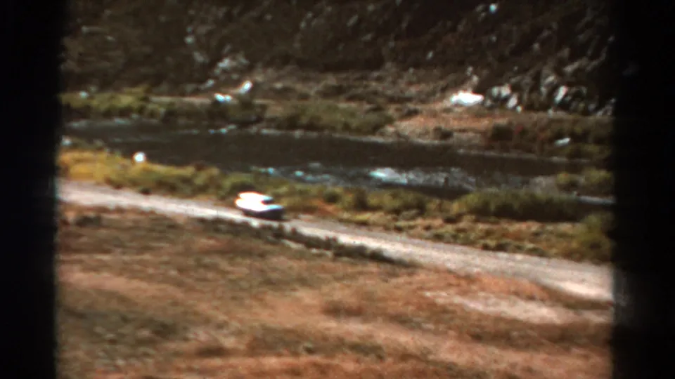 1967 Colorado home-movie frame with a white car on a pale dirt road near water, brush, and rocky slopes.