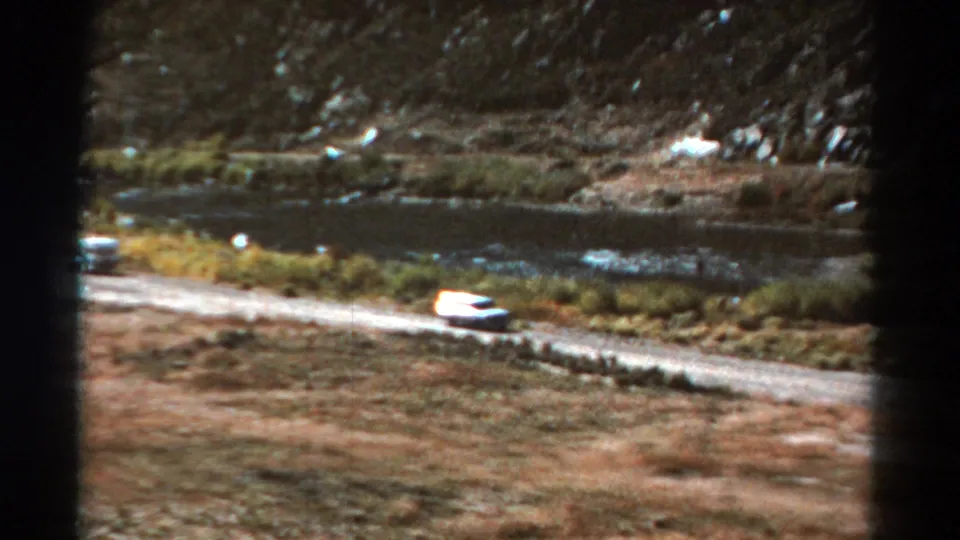 1967 Colorado 8mm frame showing the white car farther along the dirt road with water and rough hillside in the background.