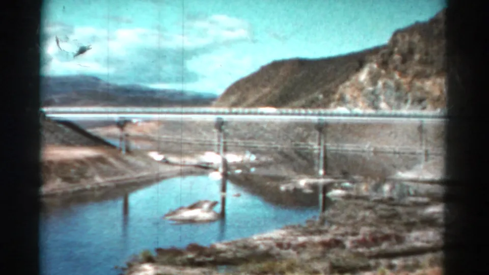1967 Colorado home-movie frame showing a bridge crossing water, with rocky hills and a pale sky beyond.