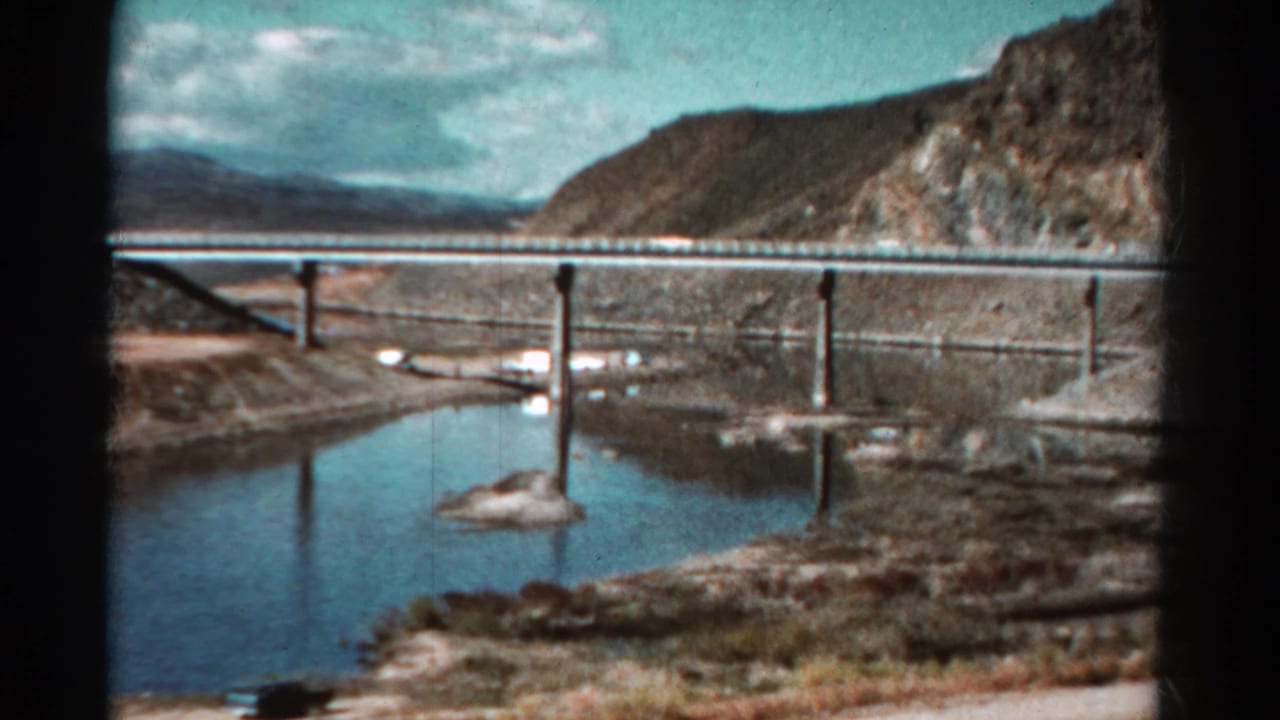 Long bridge crossing calm water with rocky hills and clouds behind it.