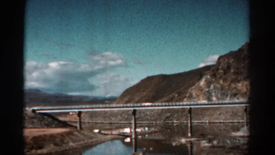 1967 Colorado 8mm frame with a bridge crossing a river or reservoir, seen from a moving car against rugged hills.