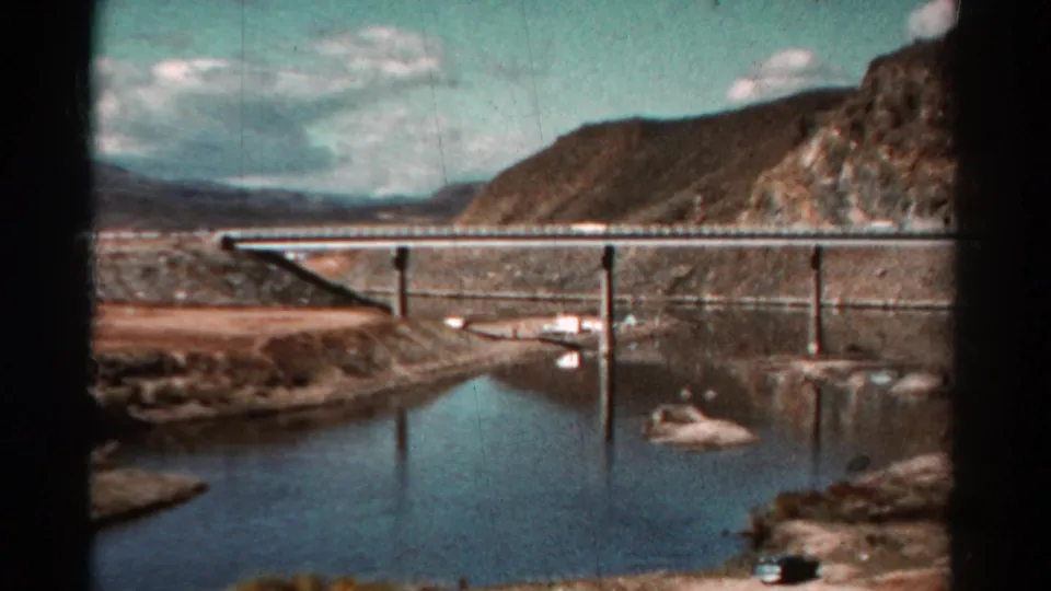 1967 Colorado 8mm frame with a wider moving-car view of a long bridge, water reflections, rugged hills, and shoreline rocks.