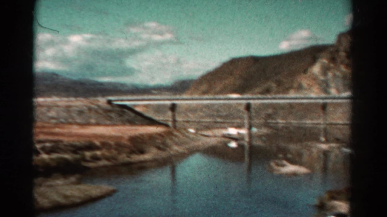 Reflective water beside a low bridge or railing with rocky hills in the background.