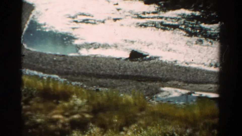 1967 Colorado 8mm frame showing grasses, gravel shoreline, bright water reflection, and a dark rock along the bank.