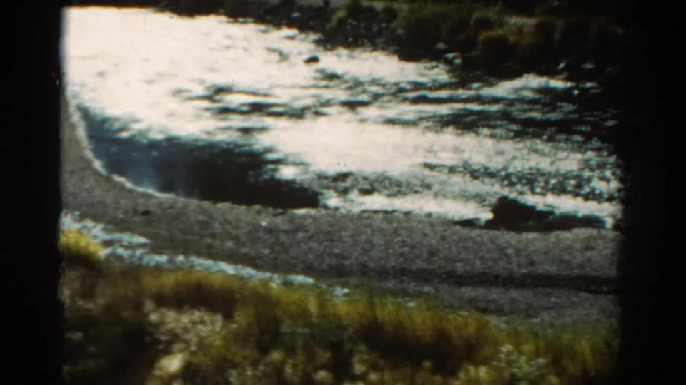1967 Colorado 8mm frame showing bright moving water, dark rocks, shoreline gravel, and roadside grass during a slow drive.