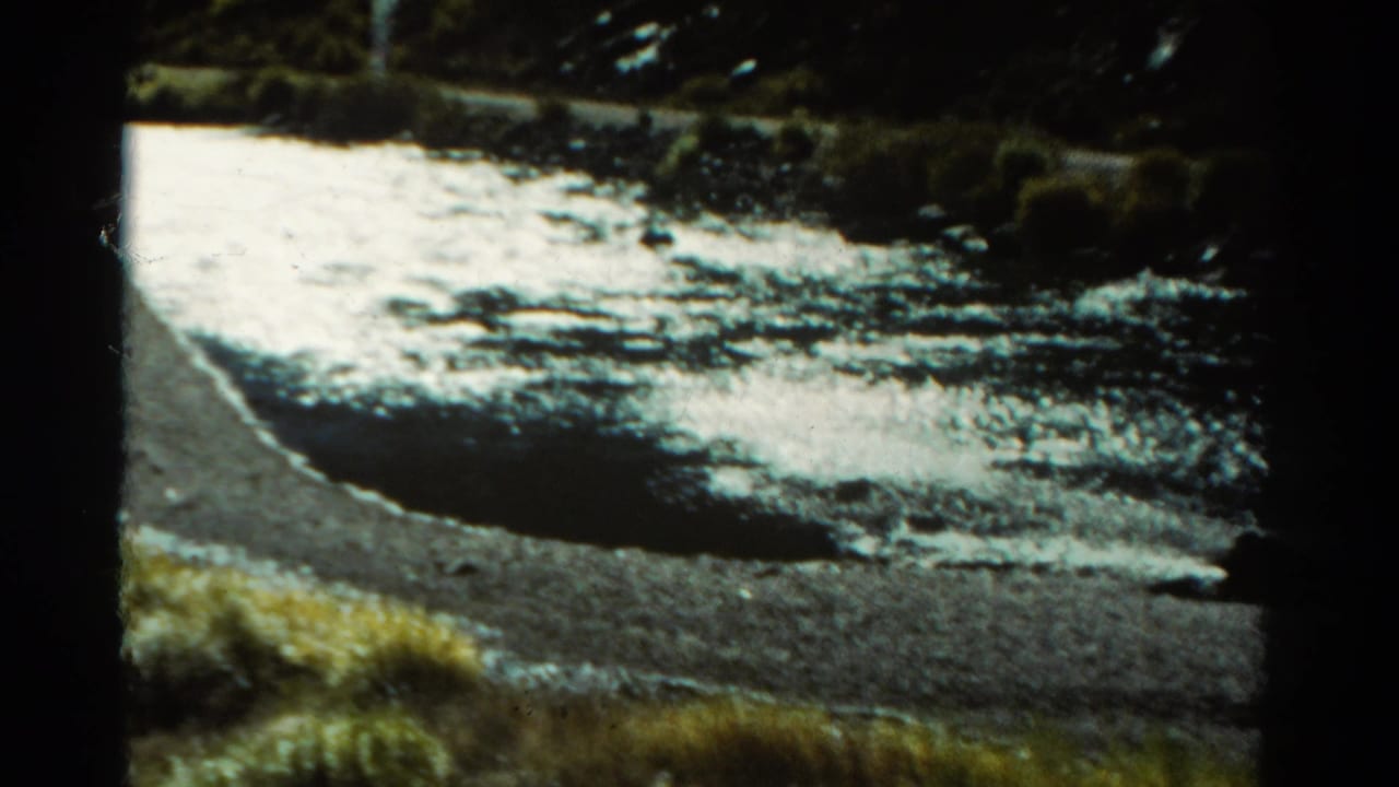 Colorado home-movie frame from 1967 with reflective white water, a curving bank, rocks, and low vegetation near the road.