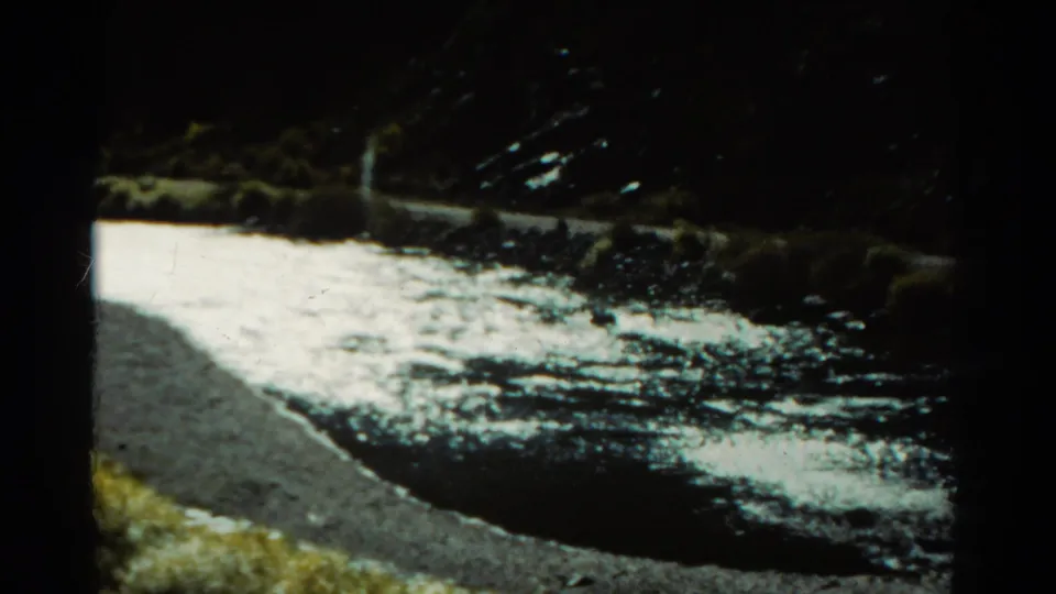Dark 8mm frame of a Colorado stream with bright water, rocky edge, grass, and heavy exposure falloff from the moving camera.