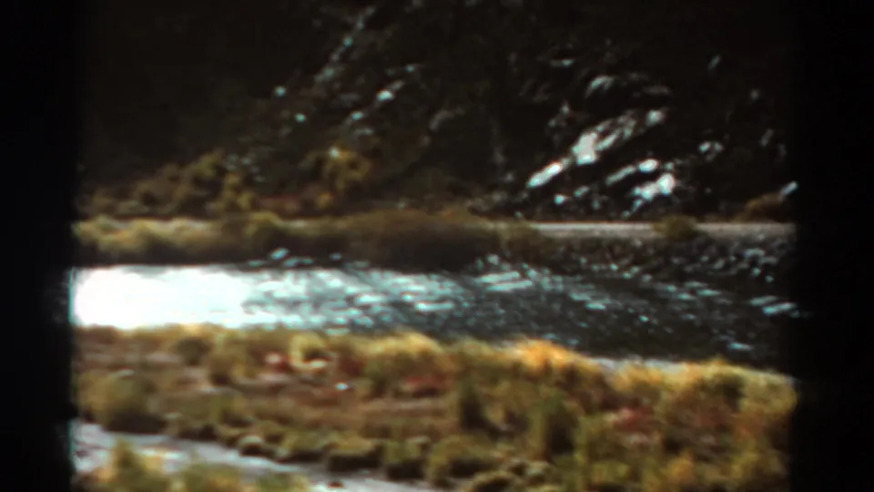 Final 1967 Colorado 8mm frame showing roadside water, sun glare, grasses, and a rocky slope from a moving car.