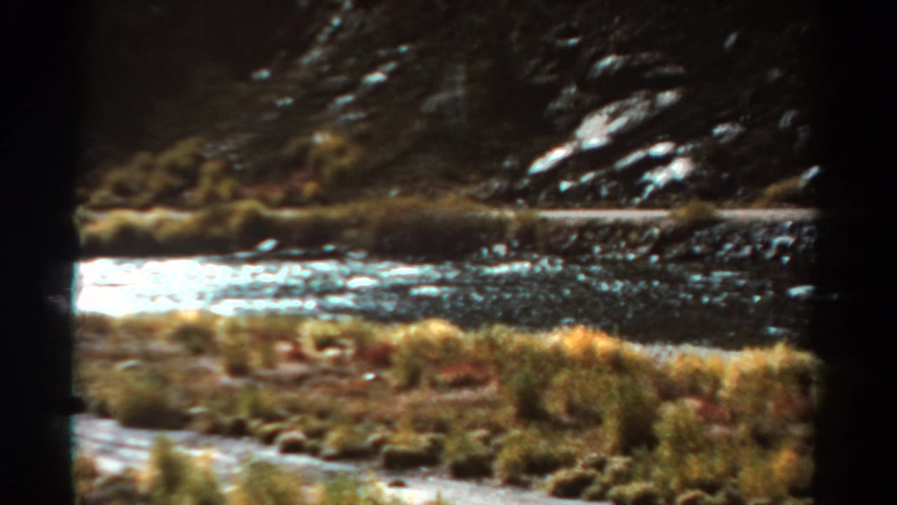 1967 Colorado 8mm frame showing a river, grassy riverside bank, and dark rocky slope from a moving travel viewpoint.