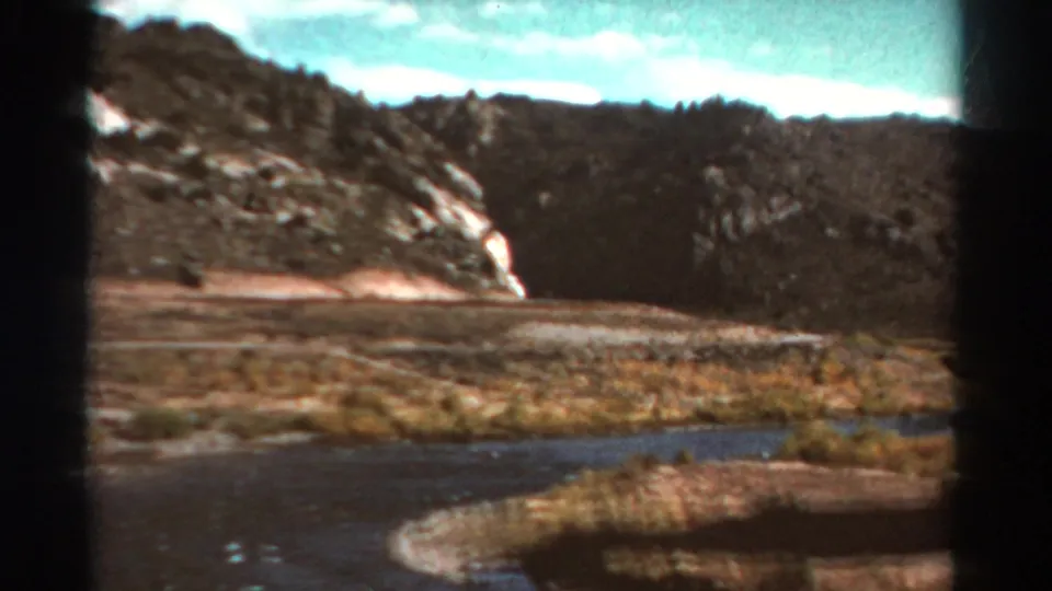 1967 Colorado 8mm view of a wider river bend, pale shoreline, rocky hills, sparse grass, and blue sky.